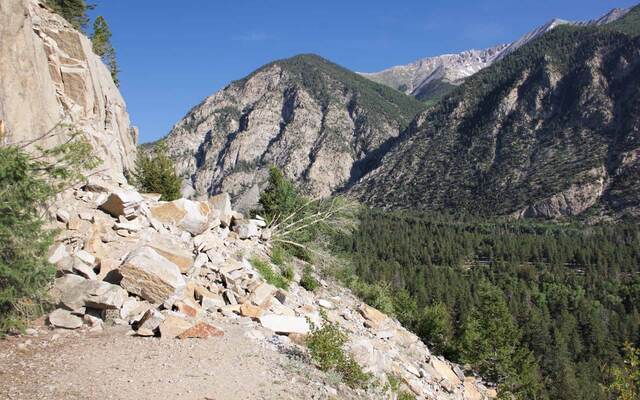 A pile of large boulders from a rockslide covering the Narrow Gauge Trail, with Chalk Creek Canyon and forested cliffs beyond