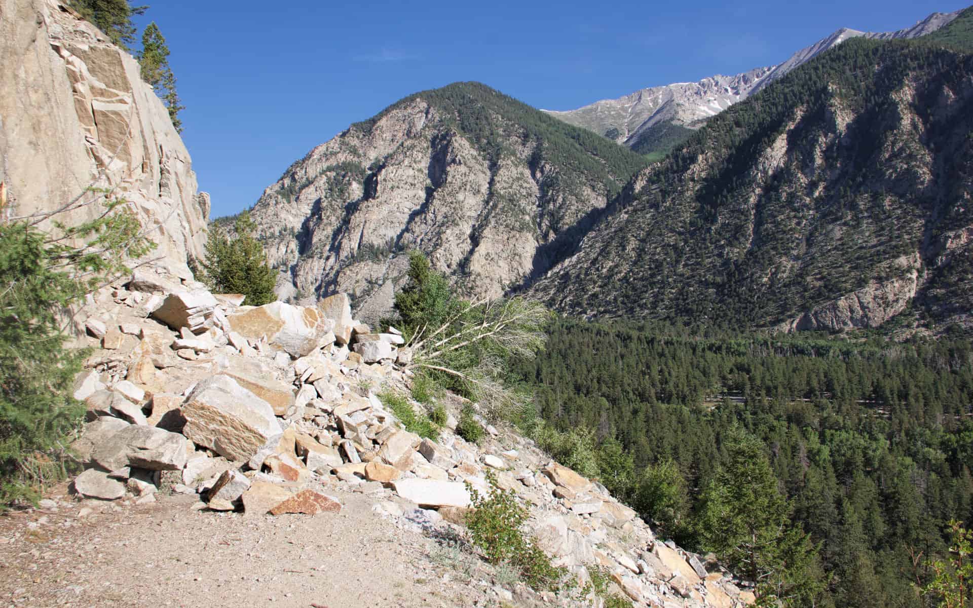 A pile of large boulders from a rockslide covering the Narrow Gauge Trail, with Chalk Creek Canyon and forested cliffs beyond