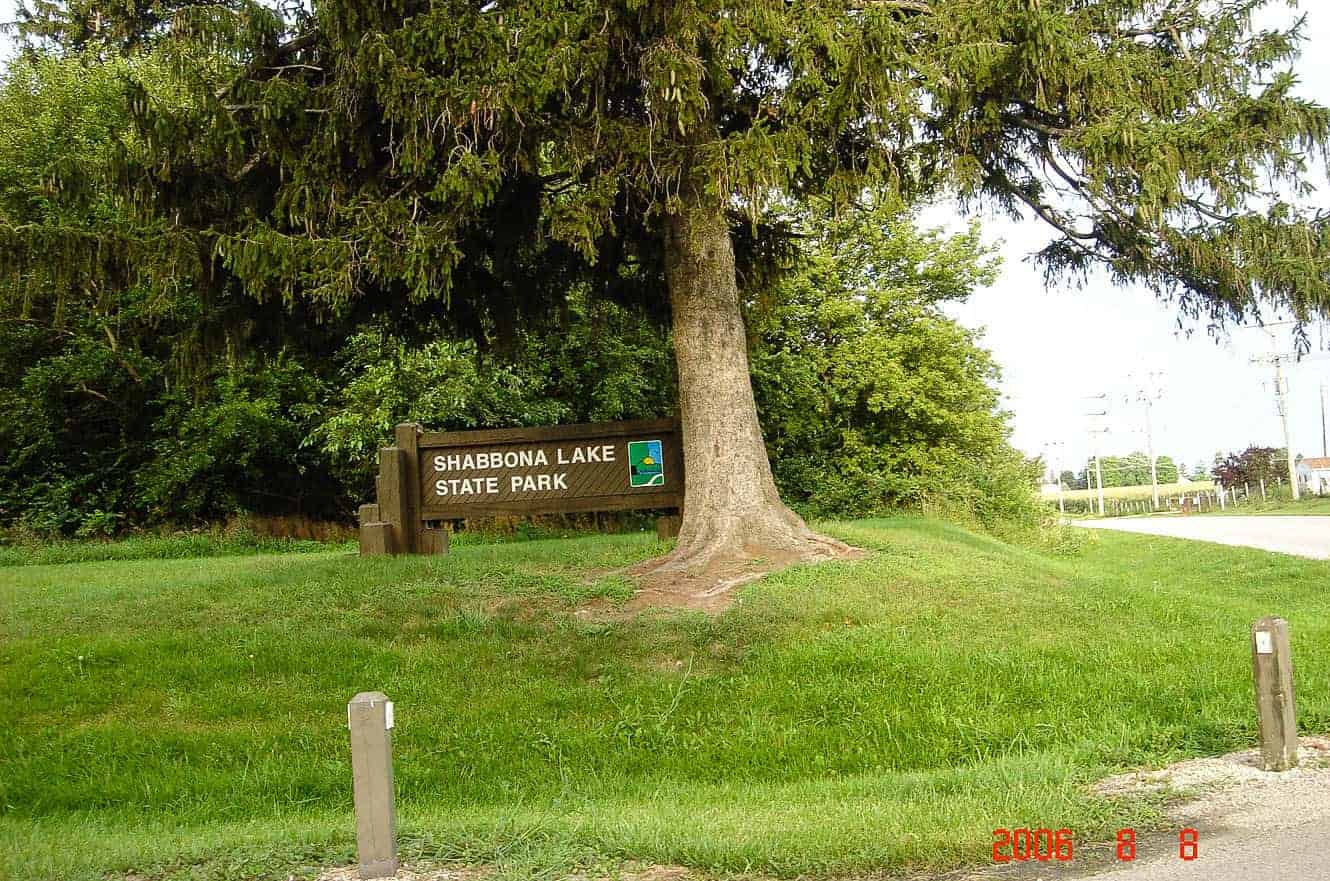 A wooden Shabbona Lake State Park sign beneath a large pine tree in Illinois
