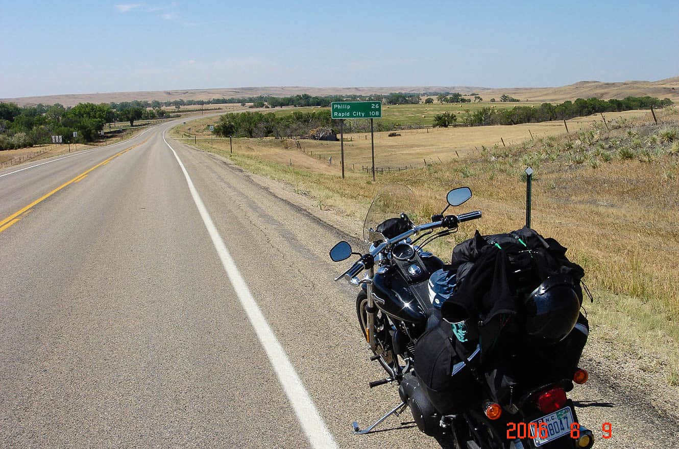 A loaded motorcycle parked roadside beside a Philip 26 / Rapid City 108 highway sign across open South Dakota grassland