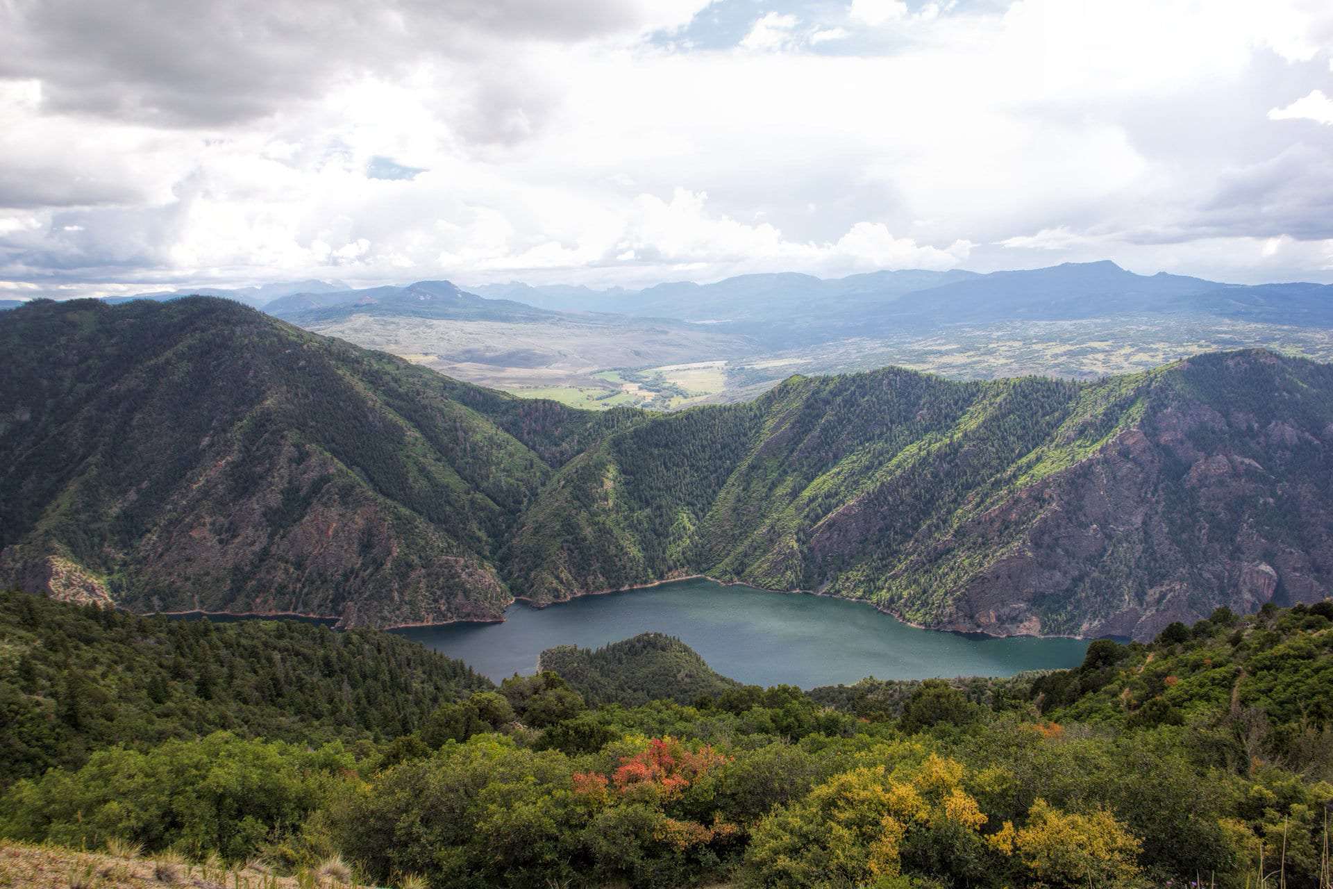 An aerial view of a reservoir surrounded by forested canyon walls, with a distant valley beyond