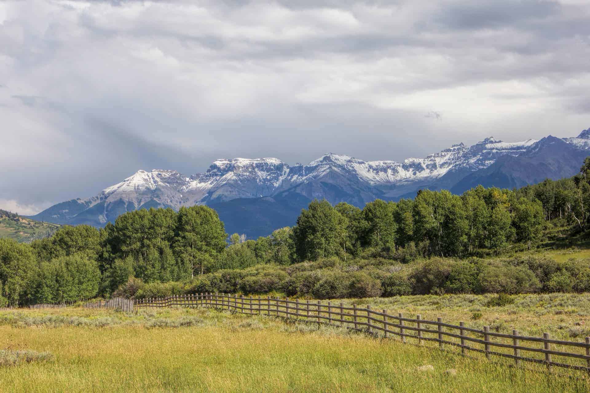 Snow-dusted peaks of the Sneffels Range rising above green ranchland and aspen groves behind a split-rail fence
