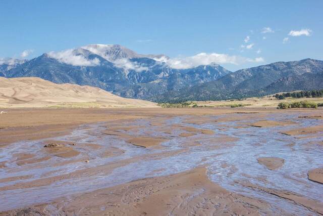 Sweeping sand dunes framed by snow-dusted peaks, with a wide sheet of shallow water and wet sand in the foreground