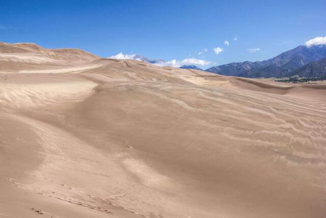 A sweeping view across rippled sand dunes toward distant mountains, with tracks crossing the dune field