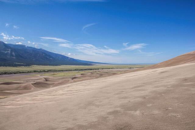 A high dune overlook across a rolling sea of sand toward a green valley and distant mountains under wispy clouds