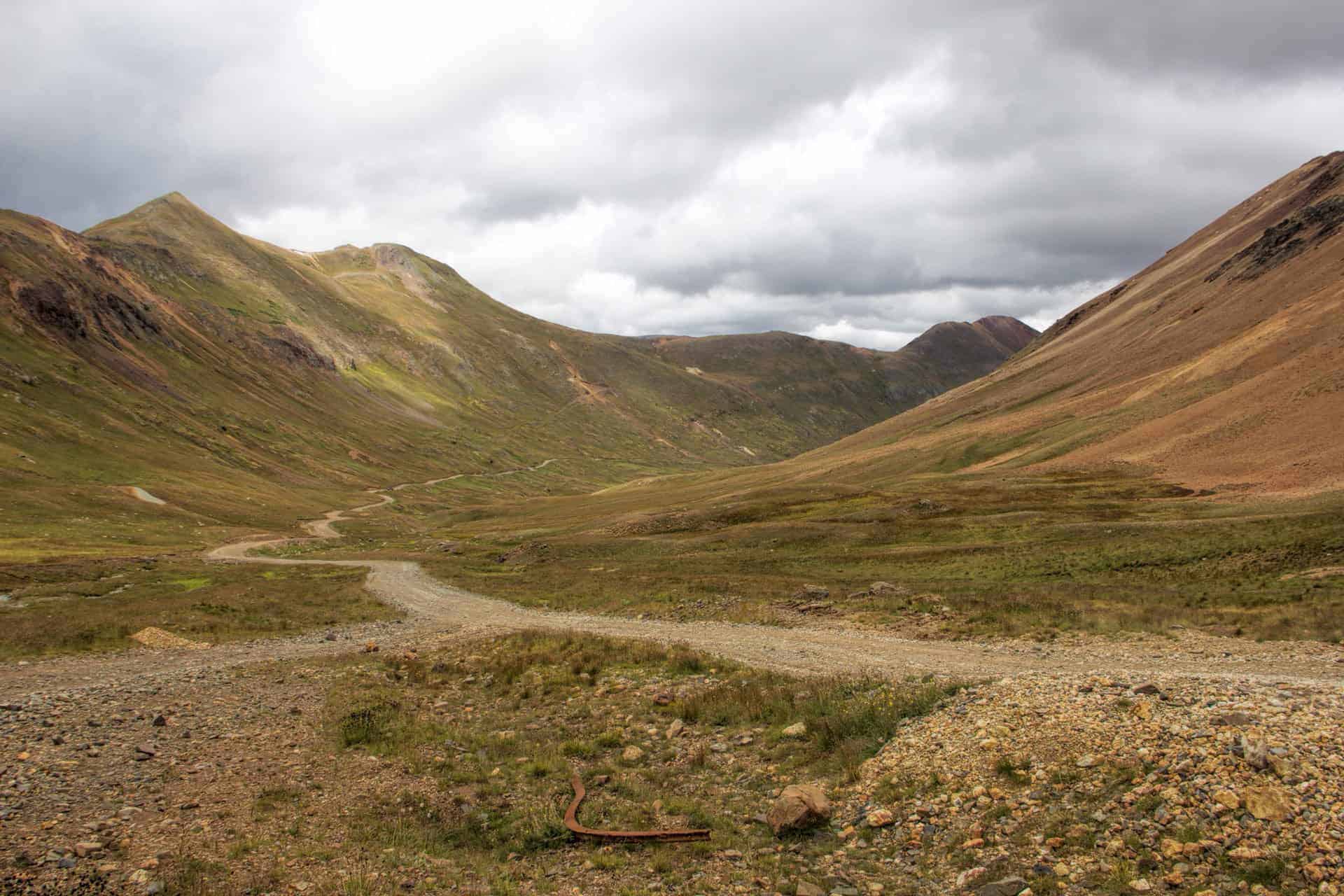 A rocky four-wheel-drive road winding through a wide alpine valley beneath layered, cloud-covered peaks