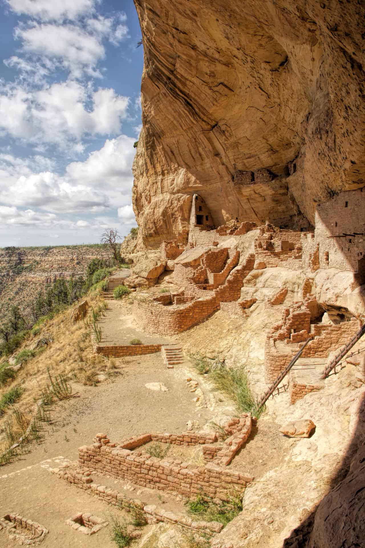 Cliff dwelling ruins tucked into a sandstone alcove, viewed from across the canyon amid pinyon-juniper forest