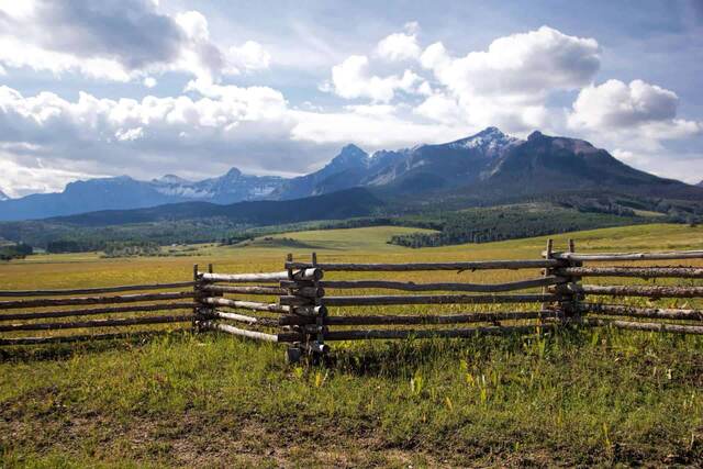 A wooden rail fence in a grassy pasture with the jagged, snow-streaked Sneffels Range in the background