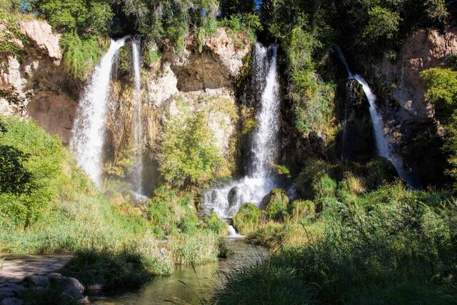 Triple waterfalls cascading over a moss-covered limestone cliff into a pool surrounded by lush greenery