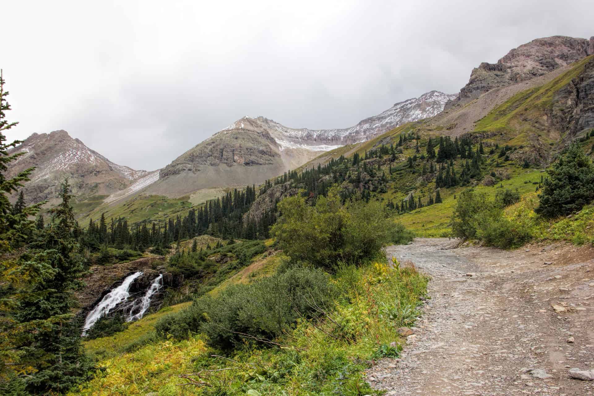 A waterfall cascading down a rocky slope beside a gravel trail leading toward cloud-covered peaks near Yankee Boy Basin