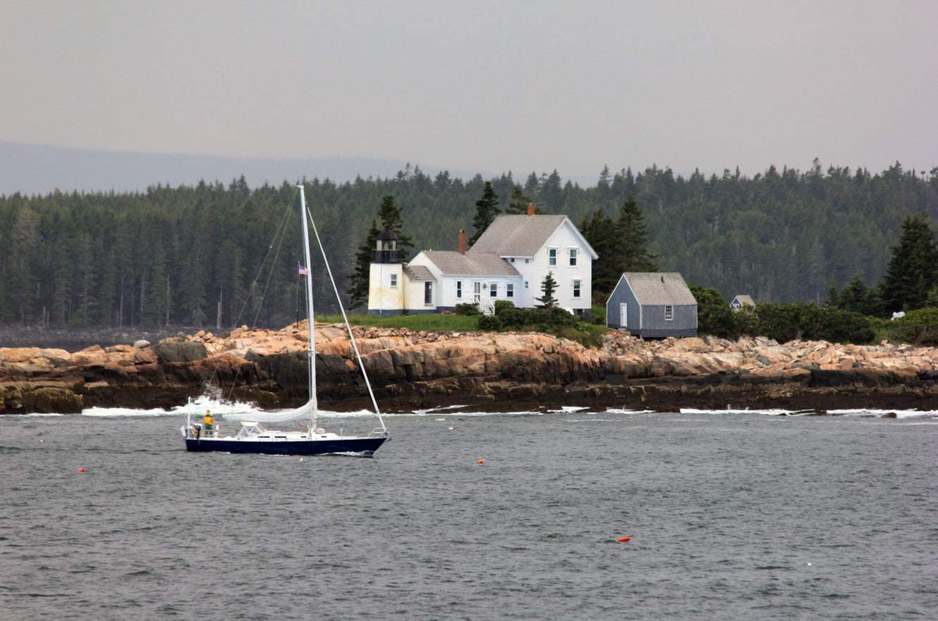 A lighthouse keeper's house on a rocky point along the Maine coast, with a sailboat passing by