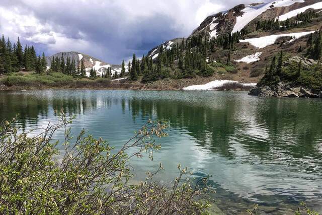 A clear blue-green alpine lake ringed by pine trees and rocky, snow-patched peaks