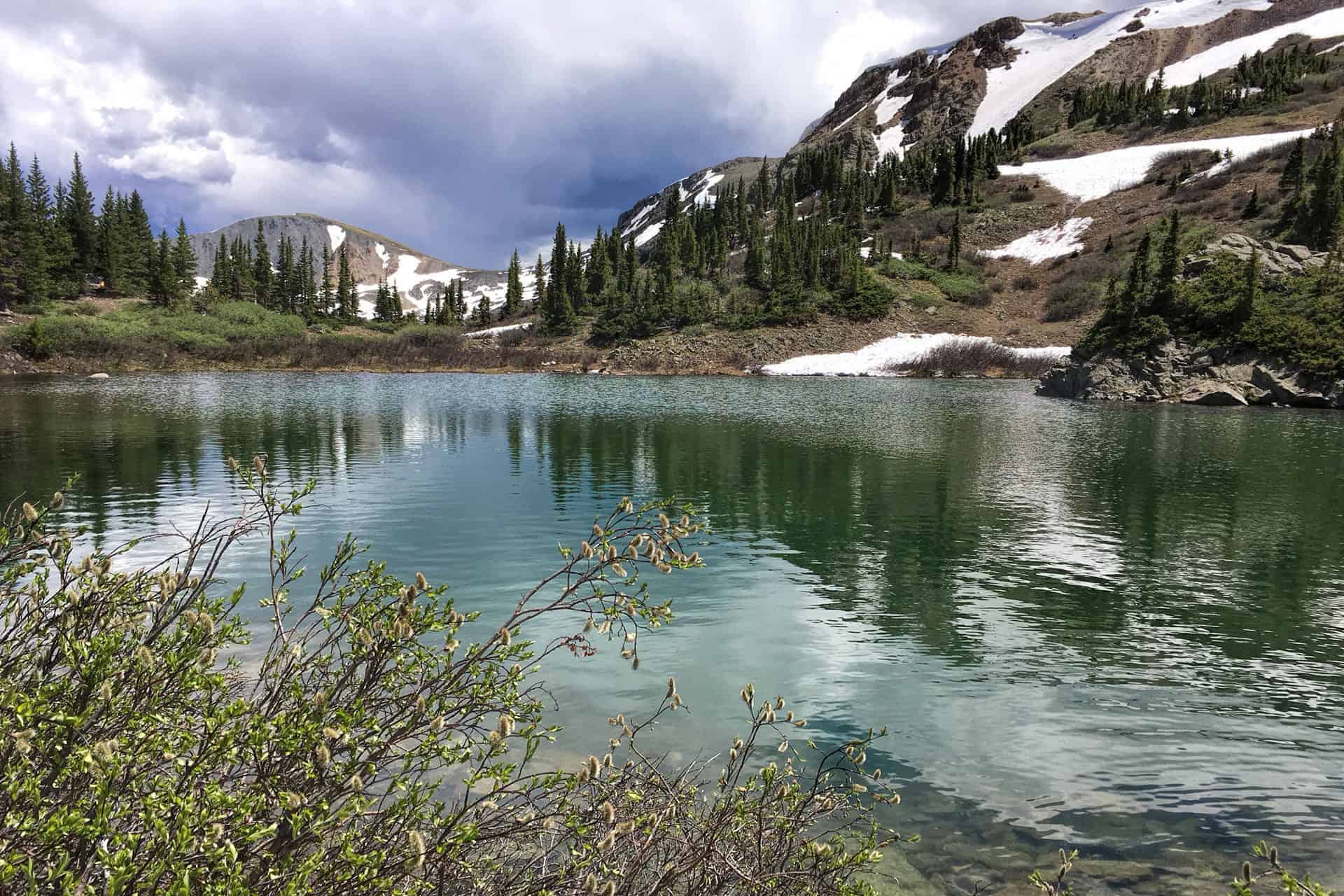 A clear blue-green alpine lake ringed by pine trees and rocky, snow-patched peaks