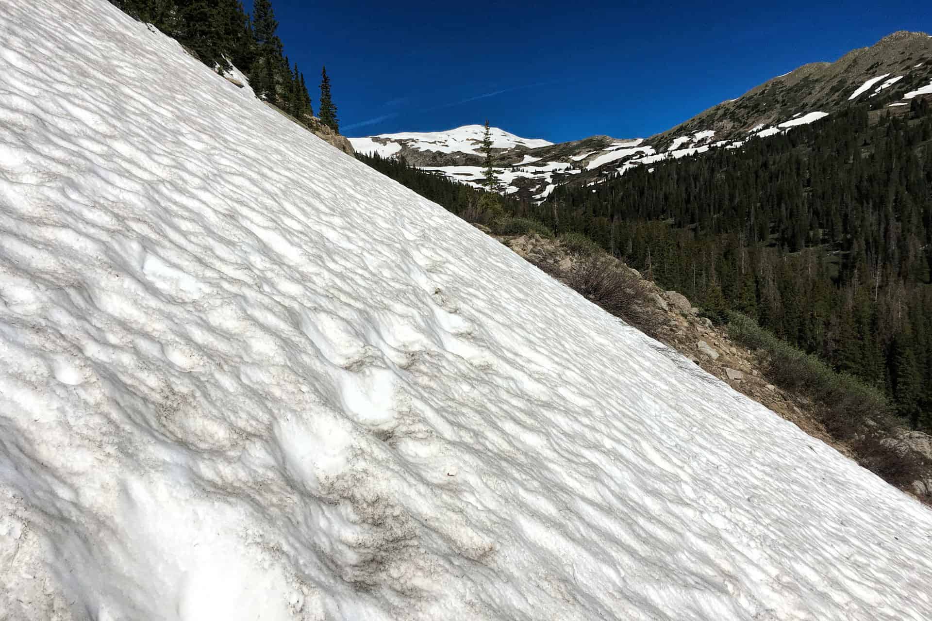 A close-up of a rippled snow slope with old footprints, framed by a distant snow-capped peak