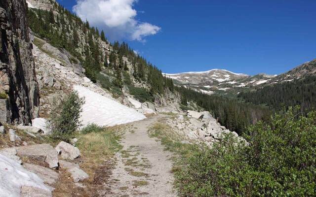 A gravel trail cutting along a rocky hillside past a lingering snowbank, with pine-covered slopes and snow-streaked peaks beyond