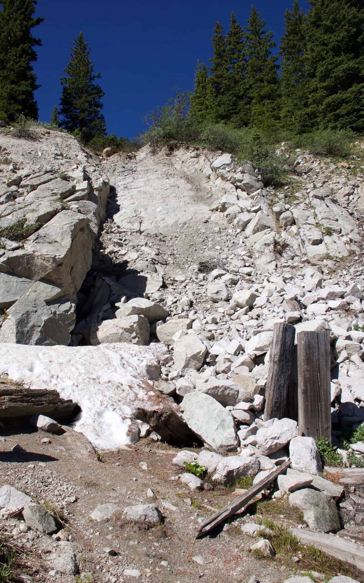 A rocky, rubble-strewn slope with a weathered wooden post and old timbers, marking the collapsed tunnel site