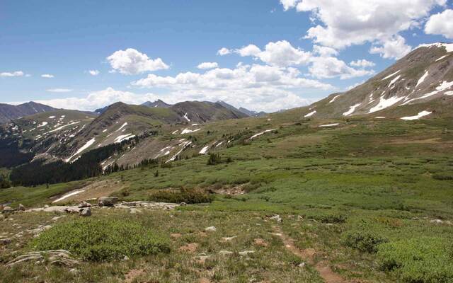 A wide mountain valley view with scattered snow patches, layered peaks, and clouds gathering on the horizon