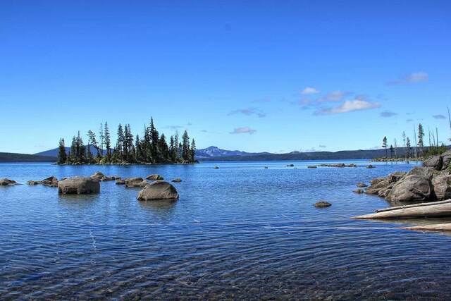 A rocky lake shoreline with a small forested island and mountains in the distance under a blue sky