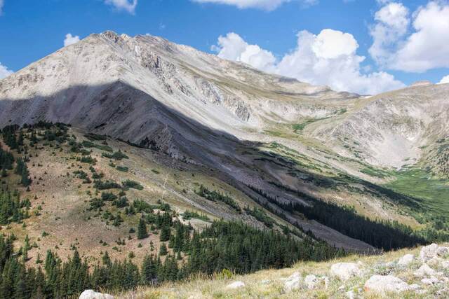 A rocky mountainside with a faint trail winding through scattered pines toward a talus slope and distant peak