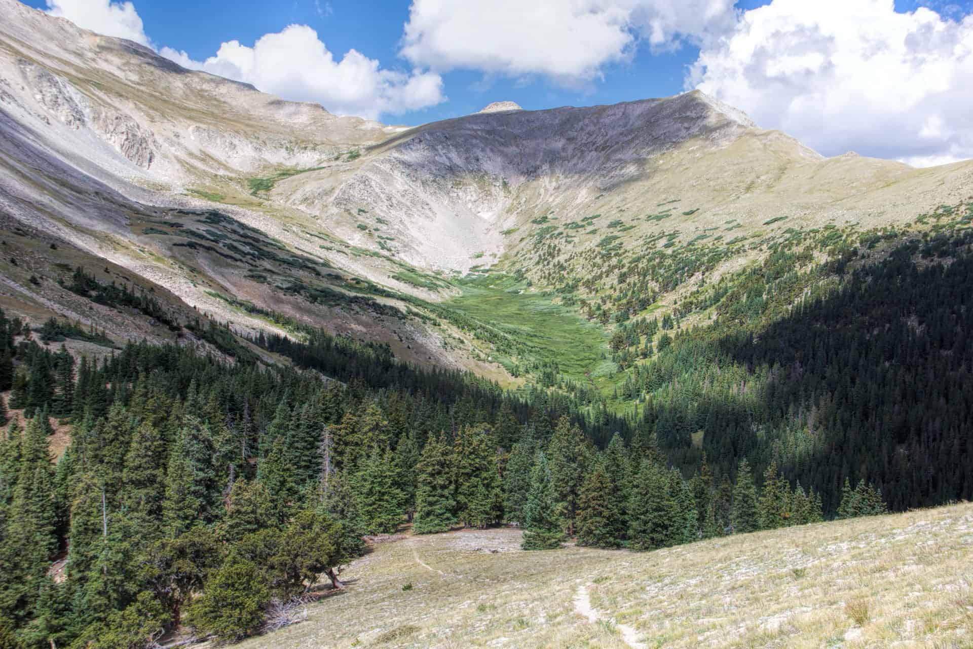A green alpine basin ringed by scree slopes and pine forest, framed by rugged peaks under a partly cloudy sky
