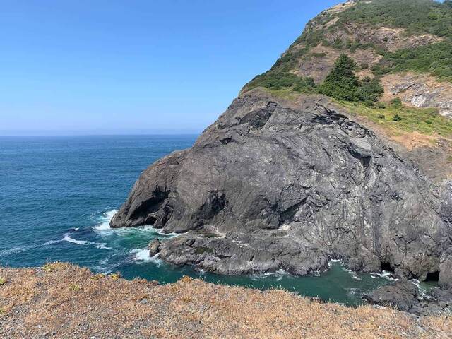 A tall rock cliff at Sisters Rock with dark sea cave openings at its base and waves rolling in