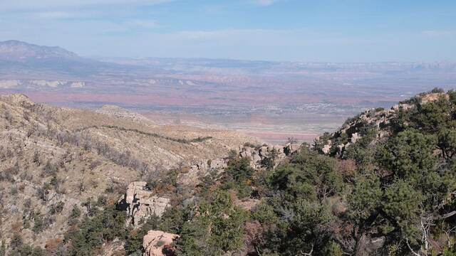 A view down a forested ridge into a valley with a small town visible in the distance, red cliff bands above