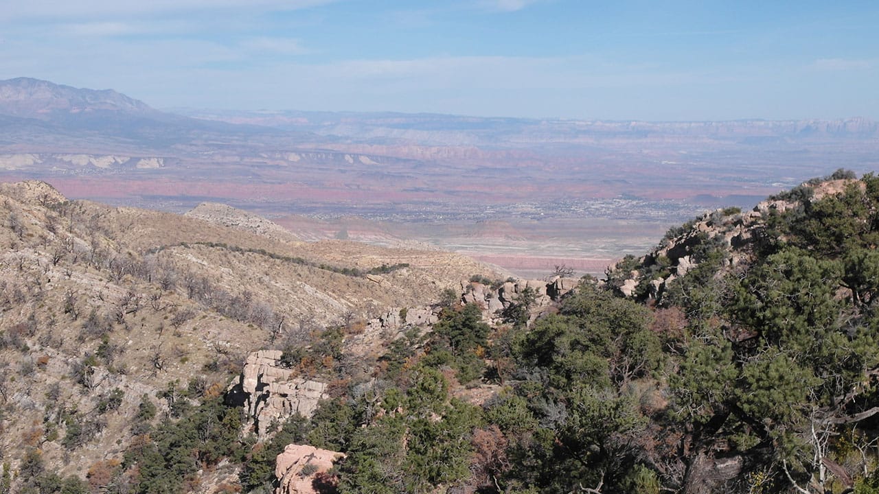 A view down a forested ridge into a valley with a small town visible in the distance, red cliff bands above