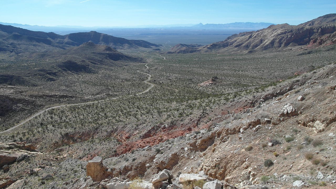 A wide desert valley with a winding dirt road, a red rock outcrop, and distant blue mountains