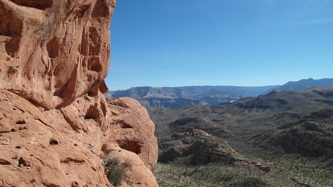 A red rock cliff face beside a sweeping view of the canyon and valley below, with a dirt road visible far down