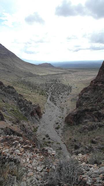 A vertical view down a rocky wash between canyon walls opening onto a wide cloudy valley
