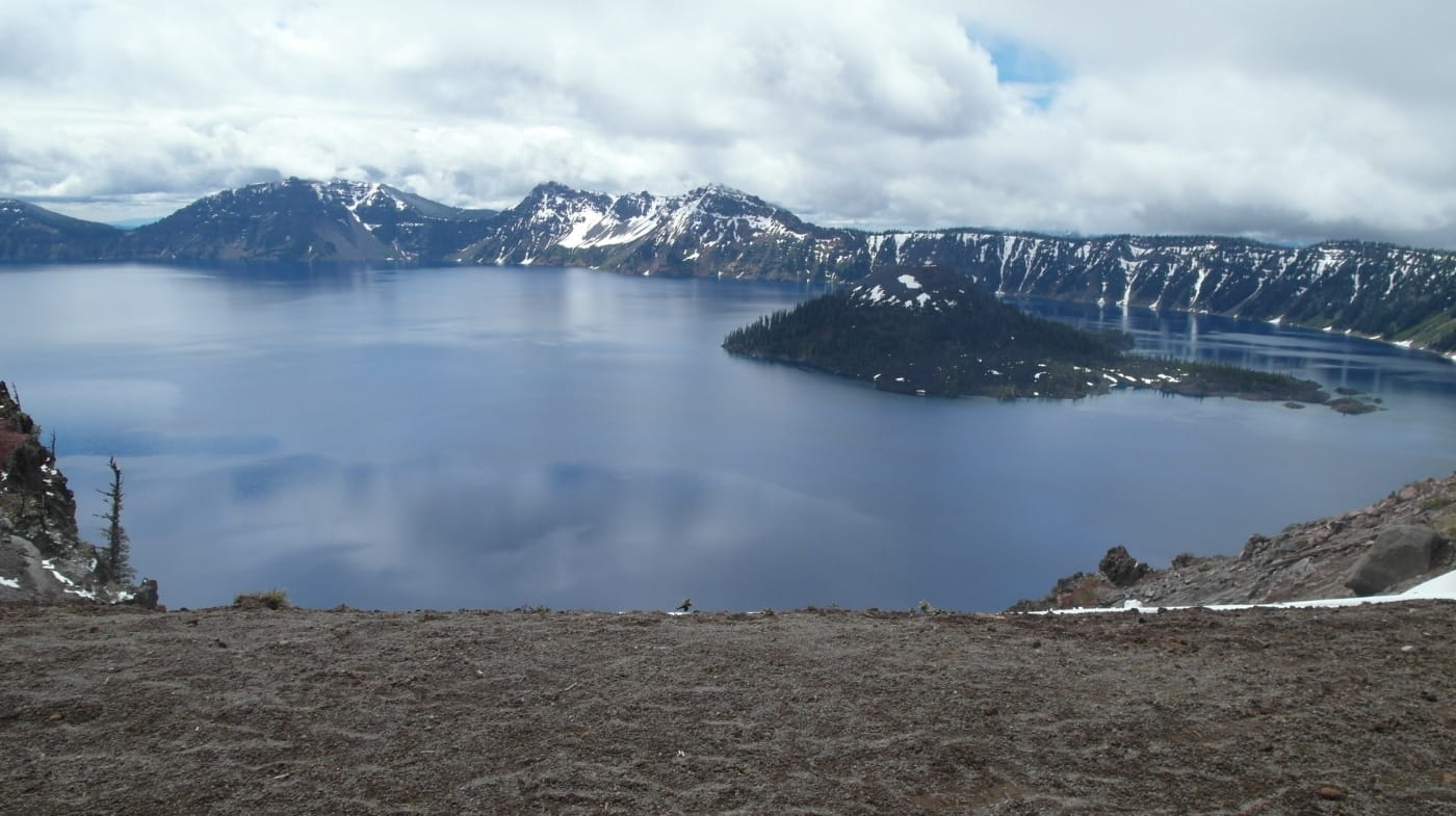 A view of Crater Lake's deep blue water and Wizard Island beneath heavy clouds and snow-streaked crater walls