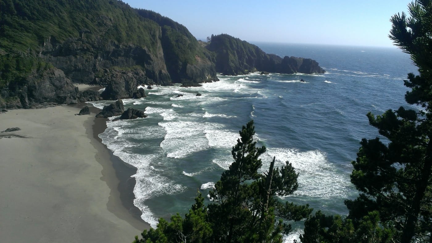 A rocky, forested headland overlooking a secluded sandy cove with waves crashing against sea stacks