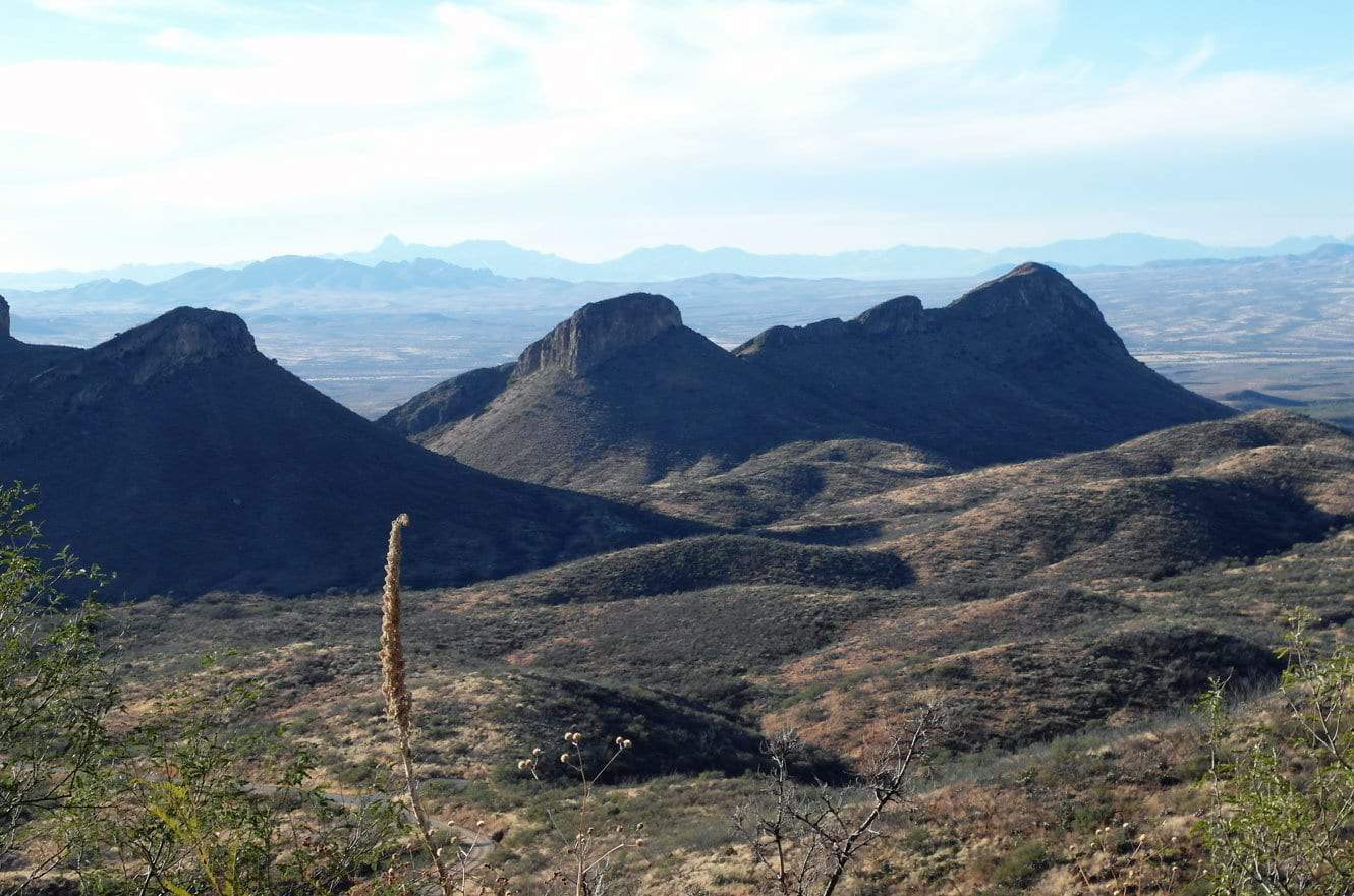 A sweeping view of layered desert mountain ridges silhouetted against a hazy horizon, with a tall agave stalk in the foreground