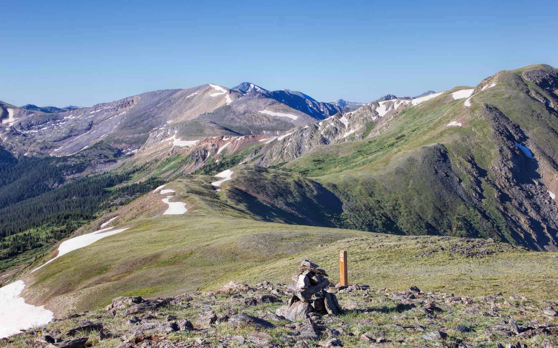 A wooden trail marker atop a rock cairn on a grassy ridge, with layered snow-patched peaks stretching into the distance