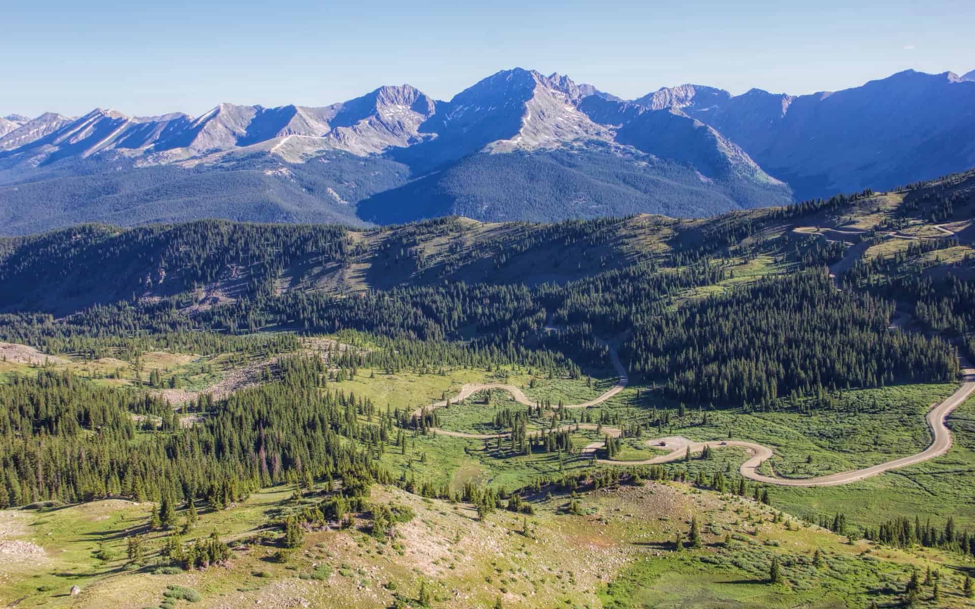 An aerial view of a winding switchback road through a green alpine valley, framed by rugged peaks