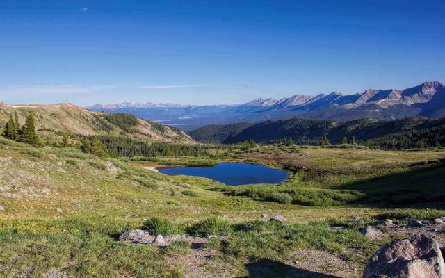 A small dark blue alpine pond in a green meadow, with forested hills and mountains beyond