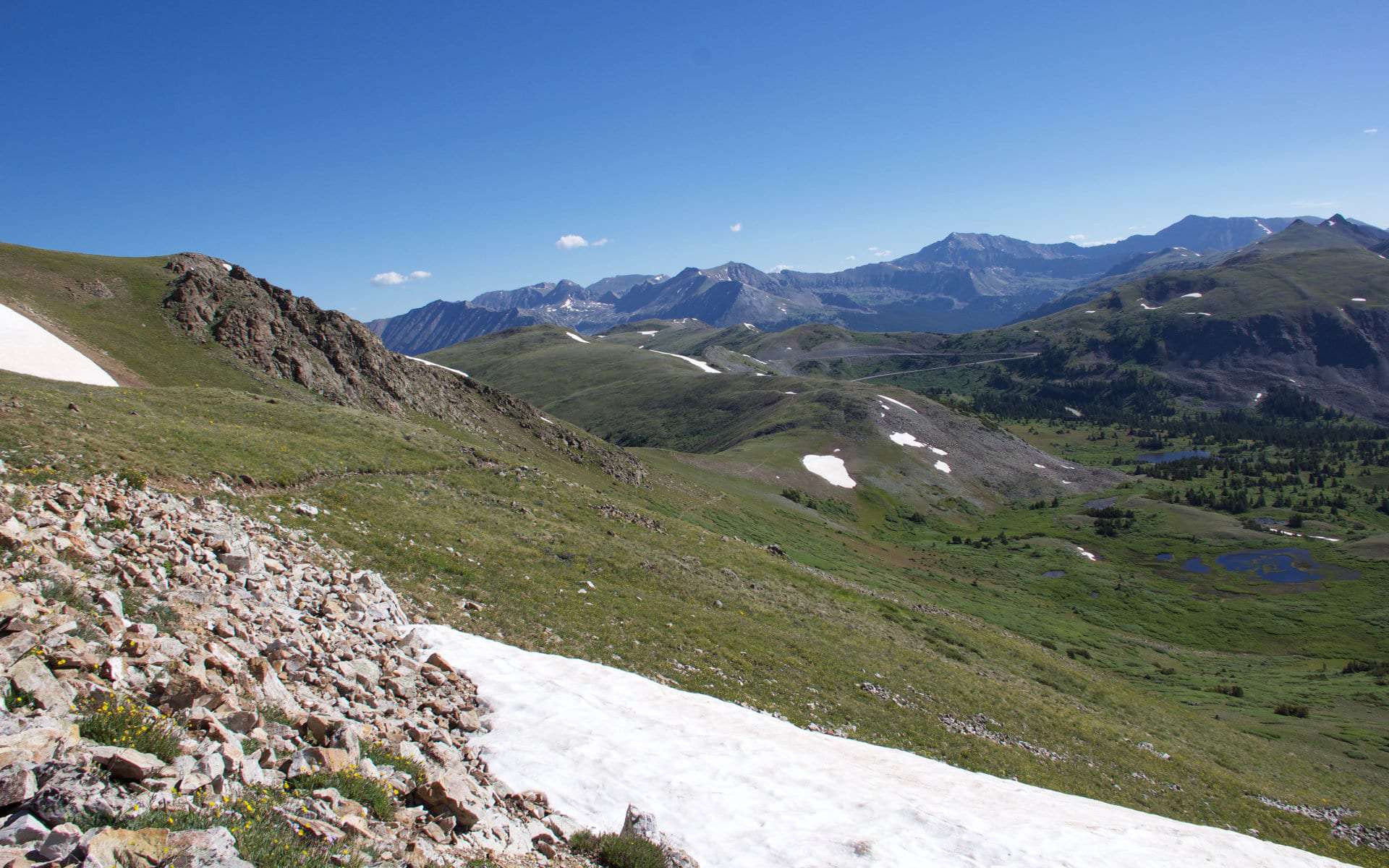 A rocky ridge trail with patches of snow, overlooking rolling green hills and distant peaks
