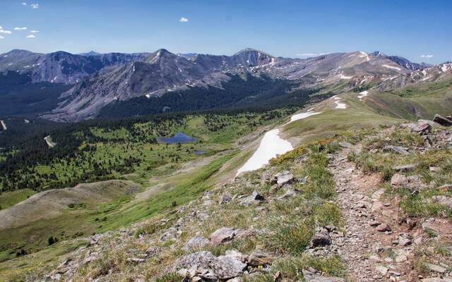 A rocky ridge trail descending toward a forested valley with a small lake, layered peaks stretching to the horizon