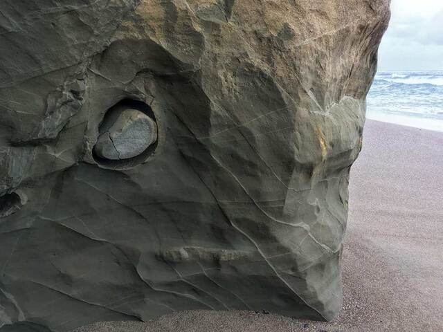 A round stone embedded in a hollow of a weathered sandstone rock face, with the ocean and surf visible beyond