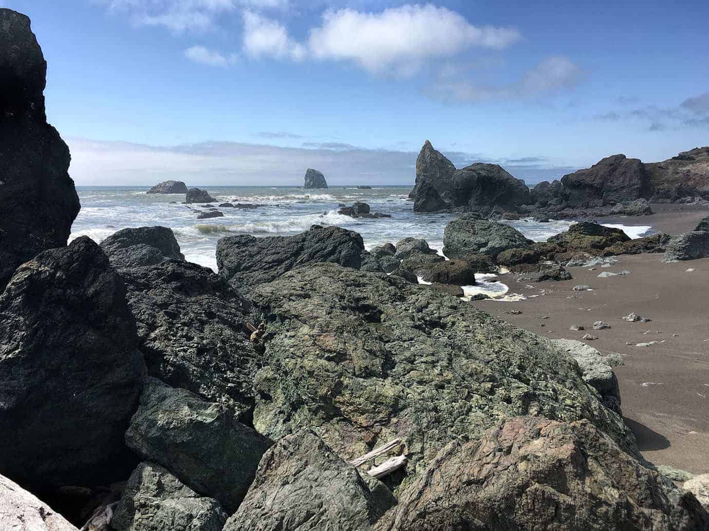 Dark, jagged rocks in the foreground with sea stacks and breaking waves along the shoreline beyond