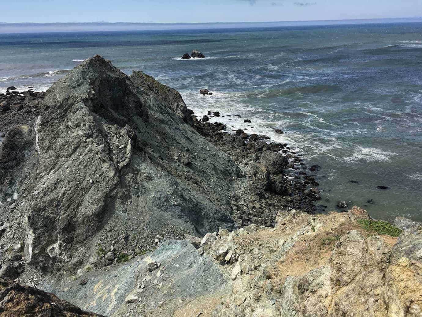 A view from a high grassy cliff edge down along the rugged coastline, with scattered sea stacks in the ocean below