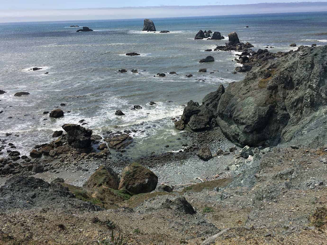 A wide view from a cliff edge across the ocean toward numerous small sea stacks scattered along the hazy coastline