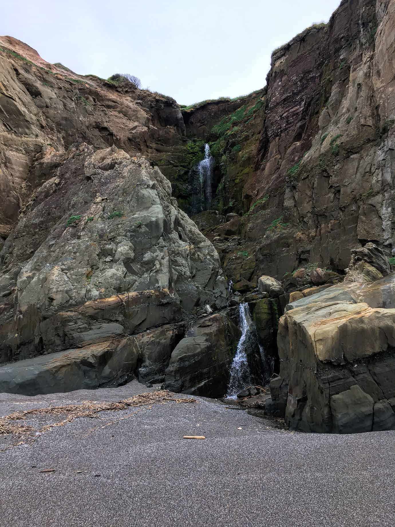 A pair of waterfalls cascading down a mossy rock cleft in the cliffside onto the beach below