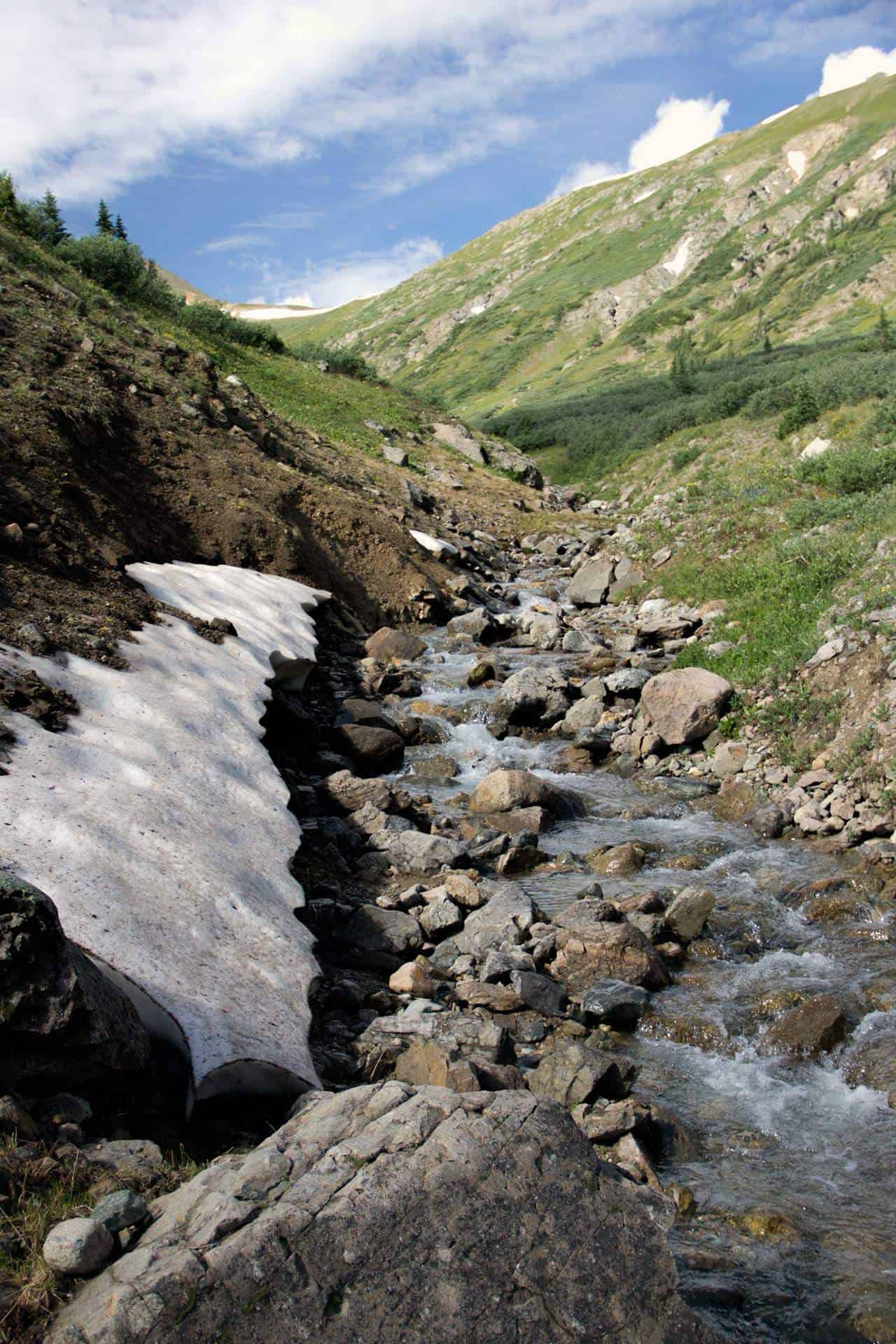 A rocky alpine stream running past a patch of lingering snow, framed by grassy slopes and distant peaks