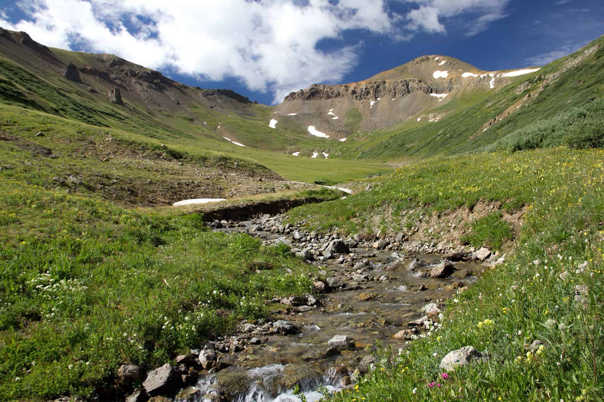 A stream winding through a green alpine basin toward a rugged, snow-streaked peak, with a faint trail visible alongside it