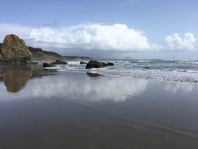 A golden sea stack rising from a calm beach with misty coastal mountains reflected in the wet sand