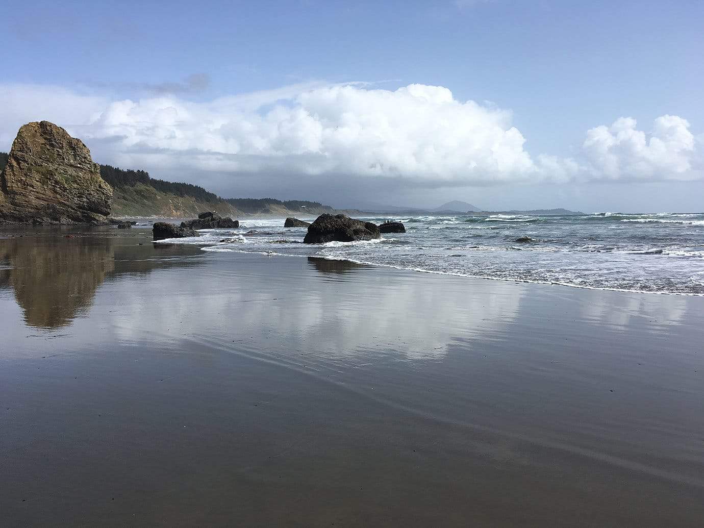 A golden sea stack rising from a calm beach with misty coastal mountains reflected in the wet sand