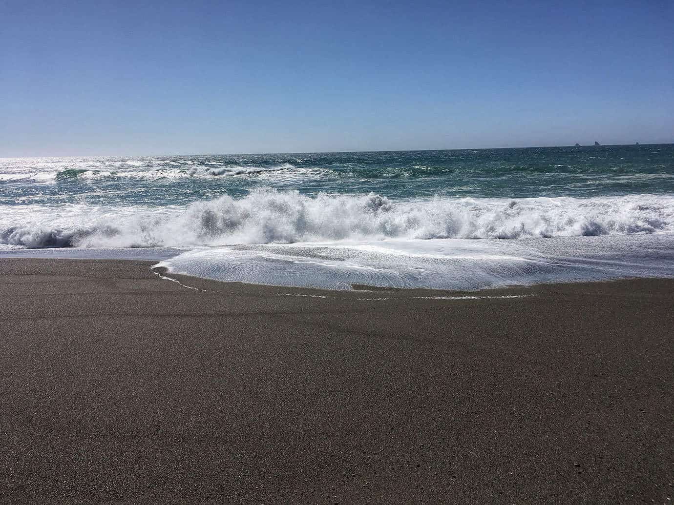 Waves crashing onto a dark sandy beach with sea stacks faintly visible on the horizon