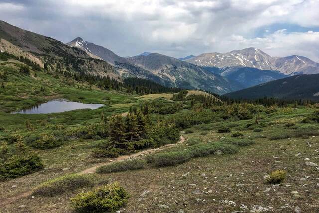 A view down a grassy alpine basin dotted with a small tarn, framed by pine trees and rugged peaks under a partly cloudy sky