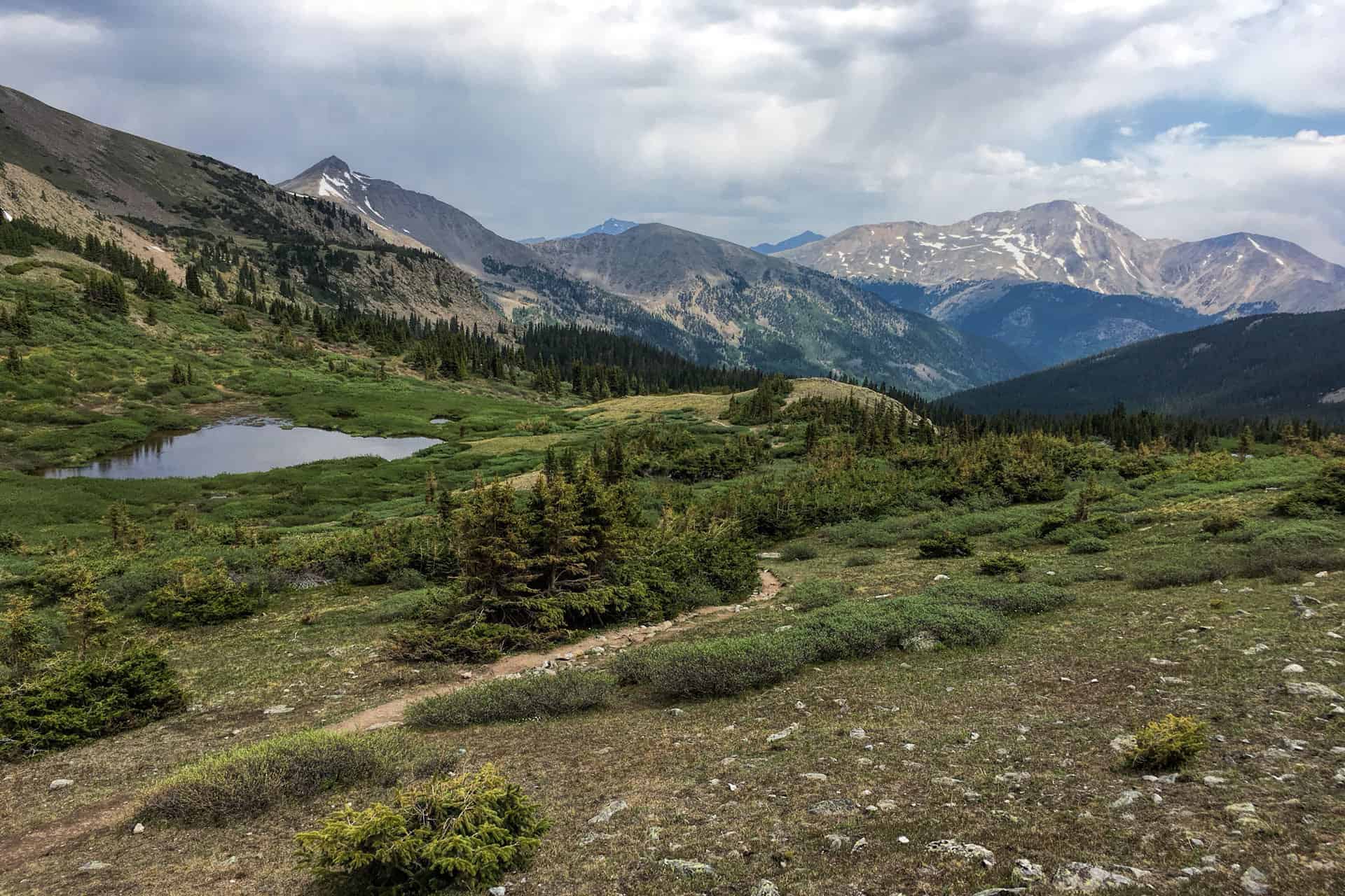 A view down a grassy alpine basin dotted with a small tarn, framed by pine trees and rugged peaks under a partly cloudy sky